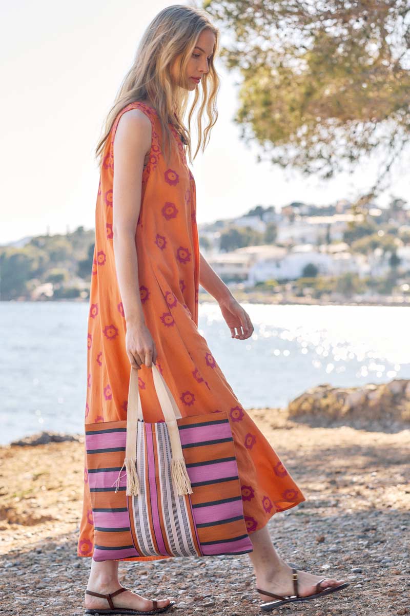 Model walks near the beach wearing East Nevaeh Sleeveless dress and carries Recycled HDPE Pink and Orange Stripe Market Bag
